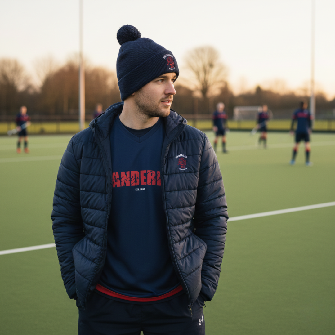 Man wearing a navy jacket and beanie on a soccer field with other players in the background.