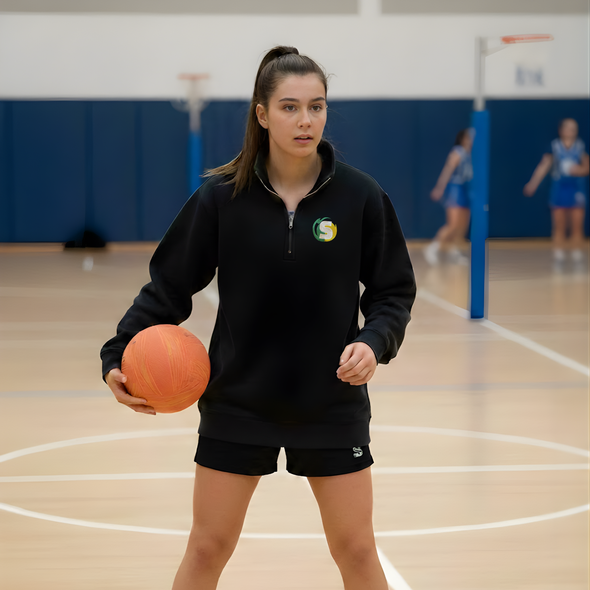 Woman holding a netball on a gymnasium floor with other people in the background