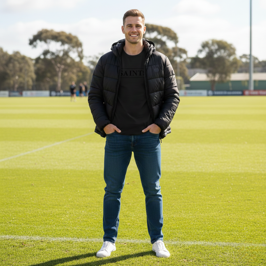Man standing on a sports field wearing a black puffer jacket and blue jeans.